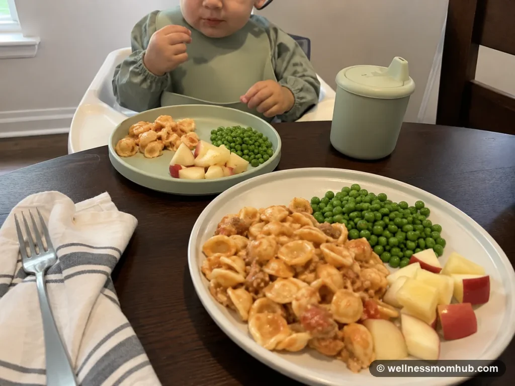 Toddler lunch ideas for a 1 year old with pasta, peas, and fruit beside an adult plate from the same meal