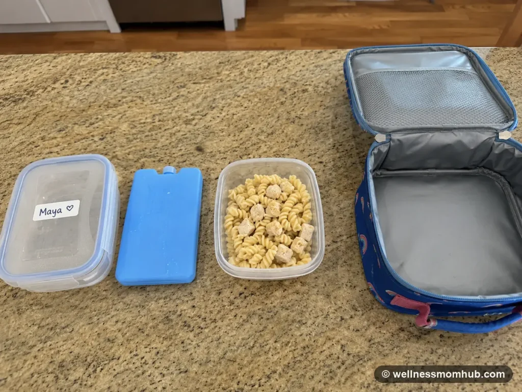 Daycare lunch packing setup with an open lunch bag, labeled container, ice pack, and a cold pasta lunch